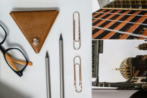 Flat lay of eyeglasses, pencils, paper clips, and leather keyholder on a white background with architectural postcard.